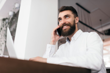 Obraz premium Successful handsome businessman using laptop computer and smartphone for his startup project, sitting in modern office. Stylish bearded man talking on mobile about business deals with partner