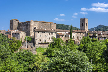 Fototapeta na wymiar Spain, Catalonia, Santa Pau: Panoramic view on the famous skyline of old ancient fortified Spanish town with tower, houses, green trees and blue sky in the background - concept travel history.