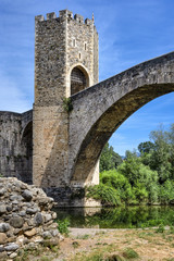 Fototapeta na wymiar Spain, Catalonia, Besalu: Panorama view on the famous skyline of old ancient fortified Spanish town with bridge, river EL Fluvia, tower, green trees and blue sky in the background - concept history.