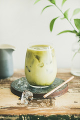 Iced matcha latte drink in glass, white wall and plant branches at background, copy space,...