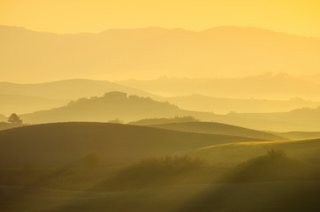 A beautiful Italian traditional rural landscape with autumn fields in the hills and a farmhouse with cypress and olive trees in a foggy morning at dawn