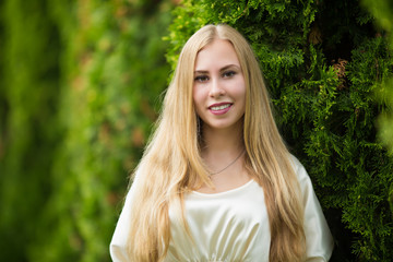 Naklejka premium Portrait of a happy young pretty blonde girl in a dress between thuja on the street in autumn.