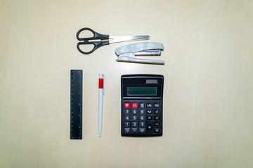 A set of office tools consisting of a calculator, pen, stapler, ruler and scissors lying on a light colored wooden table