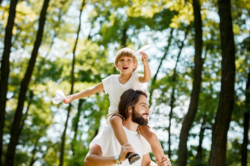 Fototapeta premium Happy boy with his father dressed in the white t-shirts walking in the park. Boy is sitting on the father's shoulders and keeps paper airplanes in his hands.