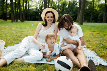 Cheerful parents with two kids dressed in white clothes is sitting on a striped blanket on the lawn on a warm sunny day in the park.