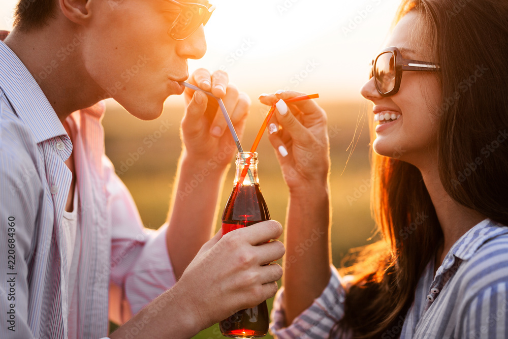 Wall mural Dark-haired girl and a young man are drinking from one bottle a drink through straws outdoor on a sunny day.