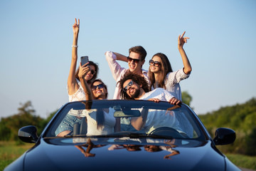 Joyful young girls and guys in sunglasses are sitting in a black cabriolet on the road holding...