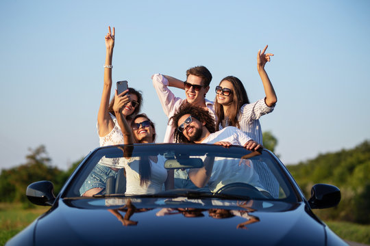 Joyful young girls and guys in sunglasses are sitting in a black cabriolet on the road holding their hands up and making selfie on a sunny day.