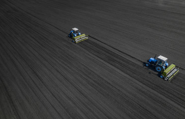 Fototapeta premium Aerial view of two blue tractors plows the earth in field on a summer day against a black earth background. Agriculture. Two tractors travel one after another along the black field
