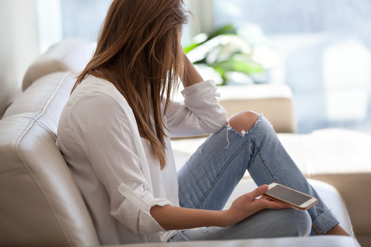 Jealous Woman Sitting On Sofa Holding Phone Feeling Sad Waiting For Call, Frustrated Millennial Girl Upset Or Worried Receiving Bad News In Mobile Message On Smartphone At Home, Cyberbullying Concept