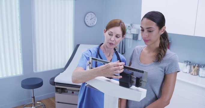Charming middle aged nurse using scale to measure weight of young latina patient