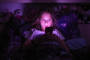 Little girl lying under a blanket looking at the smartphone at night.
