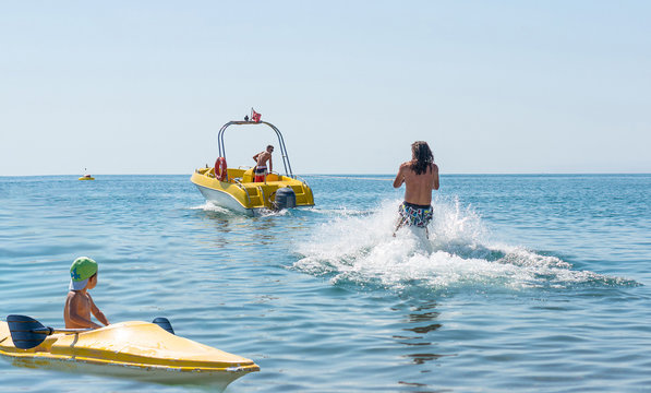 Young Man Glides On Water Skiing On The Waves On The Ocean. Healthy Lifestyle. Positive Human Emotions. Smiling Little Baby Boy In Green Baseball Cap Kayaking At Tropical Ocean Sea In The Day Time.