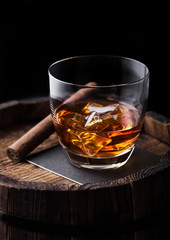 Glass of whiskey with ice cubes and cigar on top of wooden barrel. Cognac brandy drink