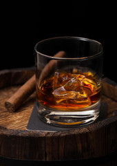 Glass of whiskey with ice cubes and cigar on top of wooden barrel. Cognac brandy drink