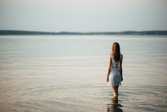 Woman in summer dress standing on seashore and looking at horizon. Young beautiful girl standing in water