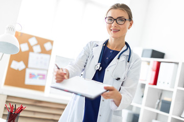 Fototapeta premium A young girl in a white coat is standing near a table in her office and holding a tablet and a pen. A stethoscope hangs around her neck.