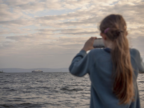Girl using her smart phone camera to take a picture of  cruise liners to share it with her friends. Ship is in focus the girl is blurred.