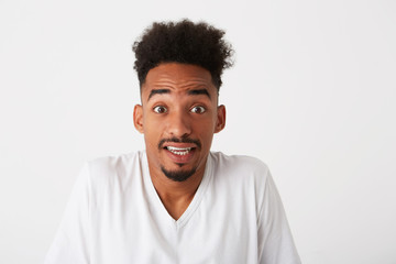 Fototapeta premium Portrait of shocked confused african american young man with curly hair wears t shirt looks embarrassed and shrugging his shoulders isolated over white background
