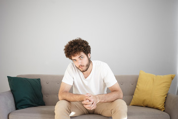Indoor shot of serious handsome young unshaven male model in white t-shirt and beige jeans posing in modern cozy living room at white wall, sitting on sofa, clasping hands and staring at camera