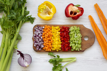 Chopped fresh vegetables (carrot, celery, red onion, peppers) arranged on cutting board on white wooden background, view from above. Top view, flat lay, overhead.