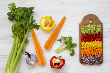 Chopped fresh vegetables (carrot, celery, red onion, peppers) arranged on cutting board on white wooden background, overhead view. From above, top view, flat lay.