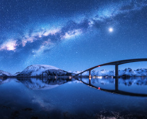 Bridge and starry sky with Milky Way over snow covered mountains reflected in water. Night landscape with road, snowy rocks, sky with moon, milky way, stars, sea. Winter in Lofoten islands, Norway 