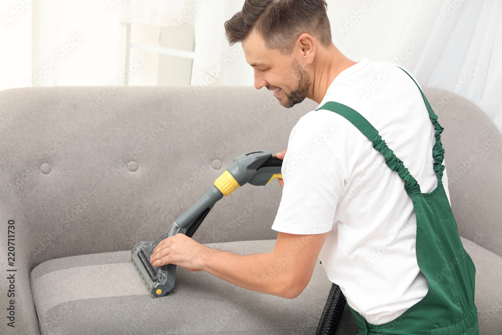 Poster male janitor removing dirt from sofa with upholstery cleaner indoors