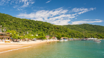 Village by the sea on an island, with a beach and a dock, people in the warm ocean water and the tropical jungle in the background