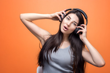 Fototapeta premium Portrait of a young beautiful girl listening to music through the headset, her hands on her head, against orange background. Lifestyle, people and technology concept