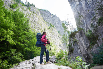 Hiker girl in modern outfit with blue backpack and trekking sticks standing before enormous stone cliff wall. Young hiker woman in pink jacket and gray pants enjoying view of high a mountain.