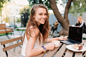 Outdoor portrait of happy smiling girl with long hair and bif eyes working outside with laptop, drinking coffee.