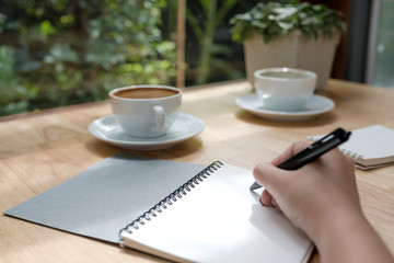 Closeup image of a woman writing down on a white blank notebook with coffee cup on wooden table