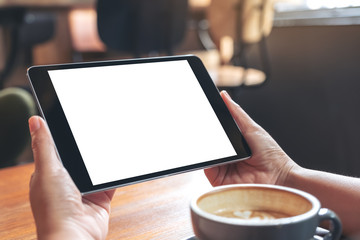Mockup image of hands holding black tablet pc with blank white screen with coffee cup on wooden table in cafe