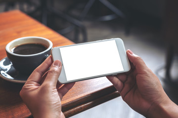 Mockup image of hands holding and using a white mobile phone with blank screen horizontally for watching with coffee cup on wooden table