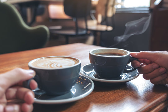 Closeup image of two people's hands holding coffee and hot chocolate cups on wooden table in cafe