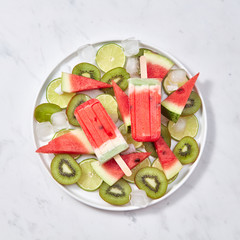 Fruit ice cream on a stick is presented in a plate with ice cubes and pieces of lime, watermelon and kiwi on a gray marble background. Space for text. Flat lay