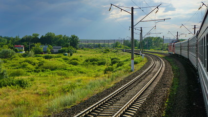 Fototapeta na wymiar Passenger train rides on rails past houses and fields on a summer evening