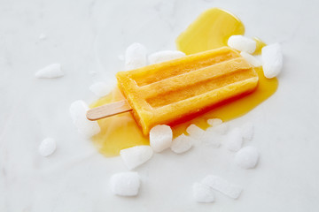 Orange fruit ice cream on a stick with slices of ice on a gray marble background with space for text. Flat lay