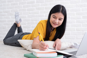 Asian woman lying on carpet and writing on notebook and learning on laptop with relax emotion at home,work at home concept.online learning.