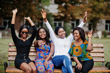 Group of four african american girls sitting on bench outdoor and put hands in the air.