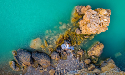 Aerial view of a young woman wearing a white dress on the rocks. Summer seascape with a girl, beach, beautiful waves, rocks, blue water. View from above. Nature