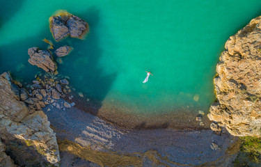 Aerial view of a young woman wearing a white dress on the rocks. Summer seascape with a girl, beach, beautiful waves, rocks, blue water. View from above. Nature