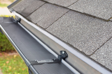 Plastic guard over new dark grey plastic rain gutter on asphalt shingles roof at shallow depth of...