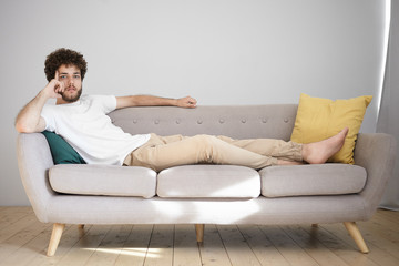 Attractive young Caucasian man with wavy hairdo and thick beard resting at home after work, lying barefooted on couch in his bachelor apartment, having pensive thoughtful expression, looking at camera