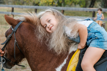 A cute little blonde girl is sitting on a pony in autumn.