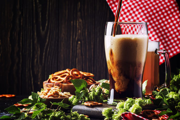 Dark German beer is poured into a glass, fresh green hops and bowls with salty snacks and nuts,...