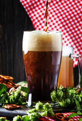 Dark German beer is poured into a glass, fresh green hops and bowls with salty snacks and nuts, autumn beer festival concept, dark background, selective focus