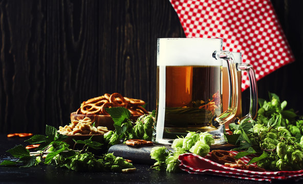Light german beer poured into big glass, fresh green hops and bowls with salty snacks and nuts, autumn beer festival concept, dark background, selective focus