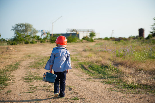 small kid wearing orange helmet with toolbox in hand walking towards factory